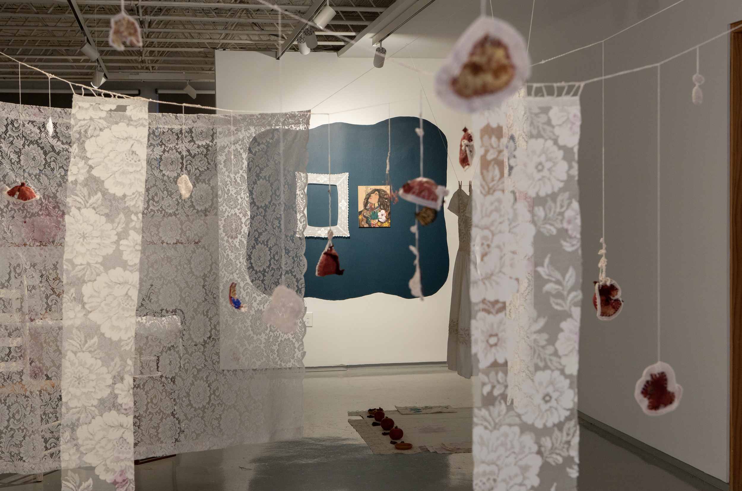 A view of Karina A. Monroy's exhibition. Lace panels and dried pomegranate skins with embroidered edges are suspended from the ceiling. Work, including a crocheted frame and a painting, can be seen hanging on the wall.