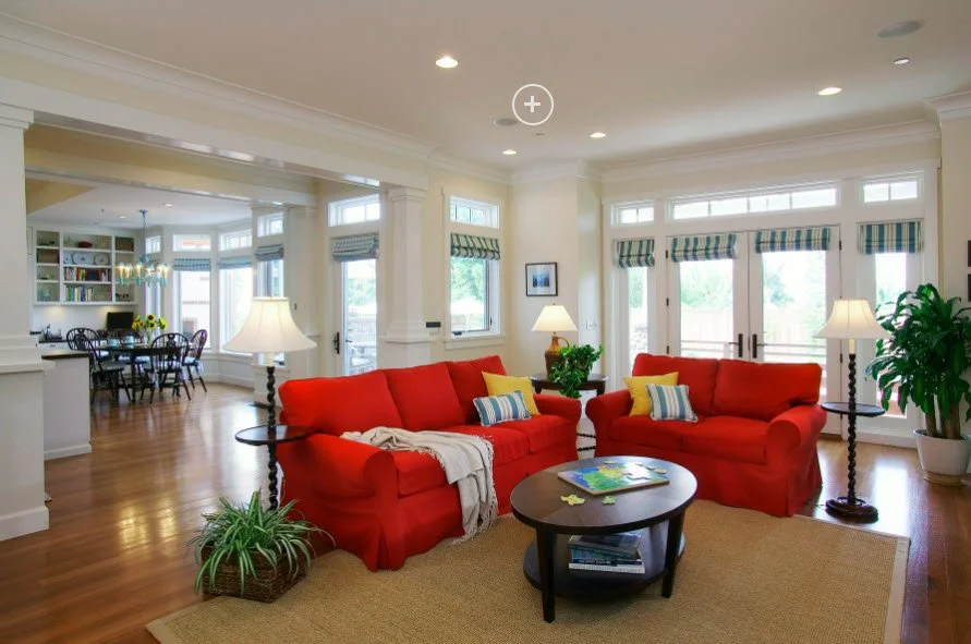 Bright living room with red sofas, wooden coffee table, throw pillows, and floor lamps. Large windows with green striped curtains provide natural light, with a dining area visible in the background.