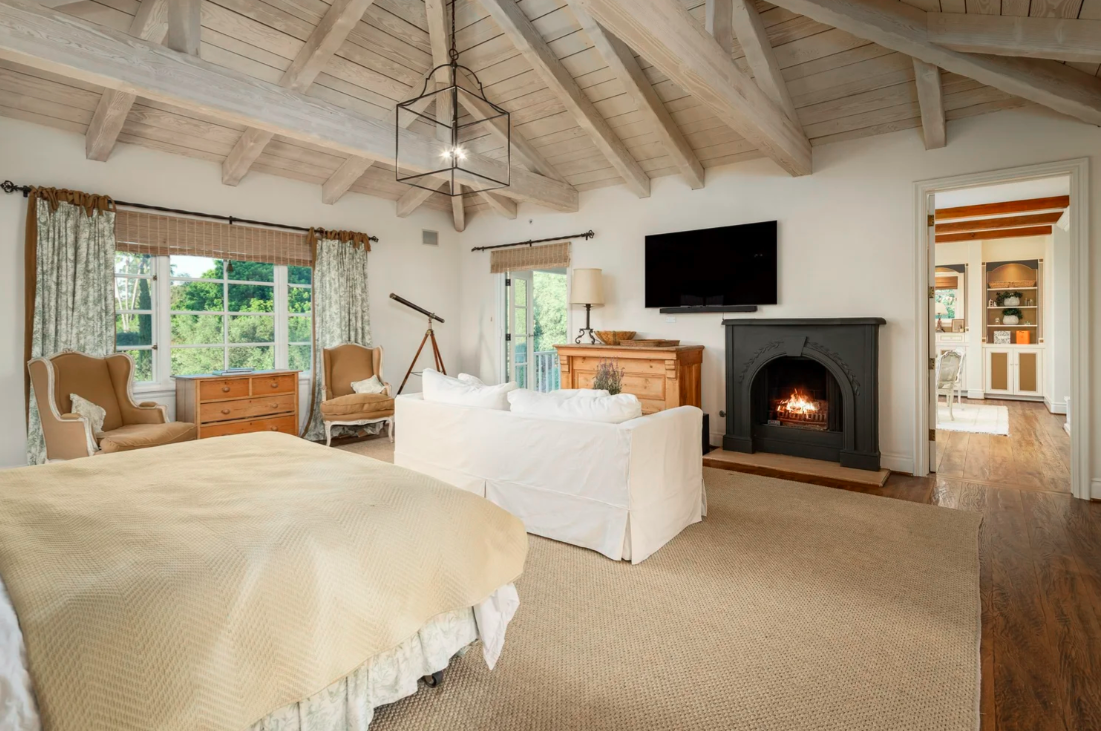A spacious bedroom with a high, vaulted wooden ceiling, large windows with floral curtains, a fireplace with a fire burning, a wall-mounted TV, a white sofa, a bed with a yellow bedspread, and classic-style armchairs.