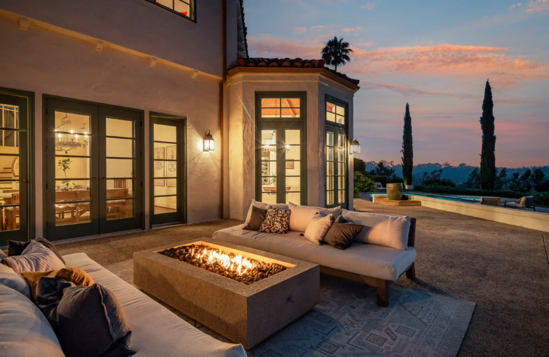Outdoor patio with sofa and fire pit at sunset, overlooking a scenic landscape with tall trees and a distant horizon.
