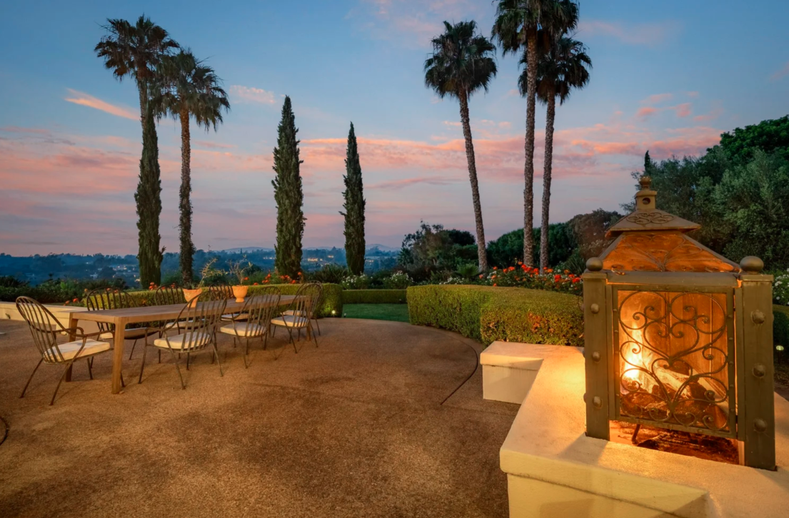 An outdoor patio area at sunset with tall palm and cypress trees, a long table with chairs, and a decorative lantern with a lit fire.