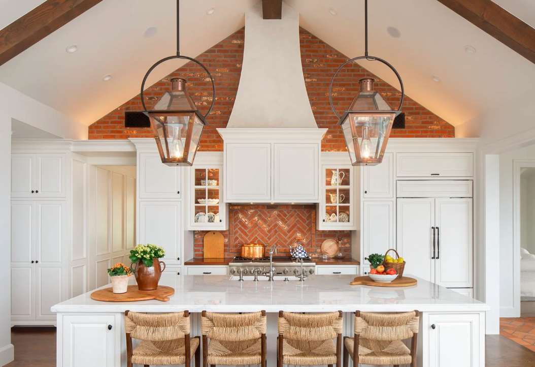 Modern kitchen with white cabinets, brick ceiling, and a large white island countertop with four woven chairs. Pendant lamps hang over the island, and decorative items like flowers, fruits, and kitchenware are on the countertop.