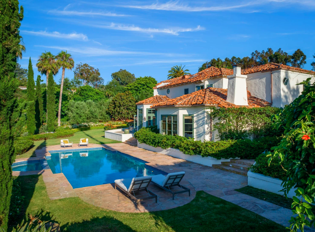 A luxurious backyard with a swimming pool, surrounded by lush green trees, a well-maintained garden, and a white house with a red-tiled roof under a blue sky.