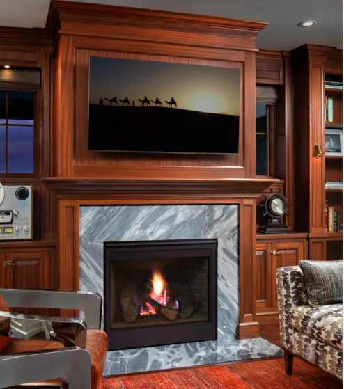 Wood-paneled living room with a fireplace, a marble surround, a mounted TV showing a desert scene with camels, and built-in shelves with books and decorative items.