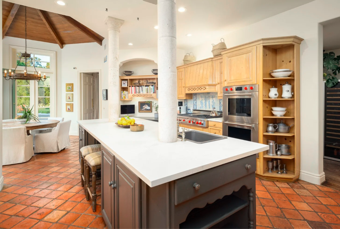 Open kitchen with white countertops, wooden cabinets, terracotta tile floor, and a breakfast bar with two stools. There's a dining area with a round table and four chairs, large windows, and decorative jars and bowls on shelves.