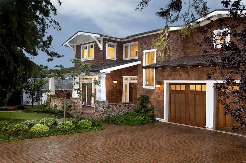 A two-story house with a brick exterior, wooden garage door, and lit windows seen during dusk, surrounded by a landscaped yard with green bushes, trees, and a brick driveway.