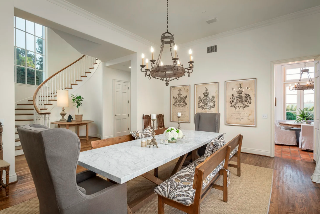A dining room with a marble dining table, upholstered and wooden chairs, a chandelier, hardwood floors, and decorative wall art. There's a staircase and a small side table with a lamp and plant.