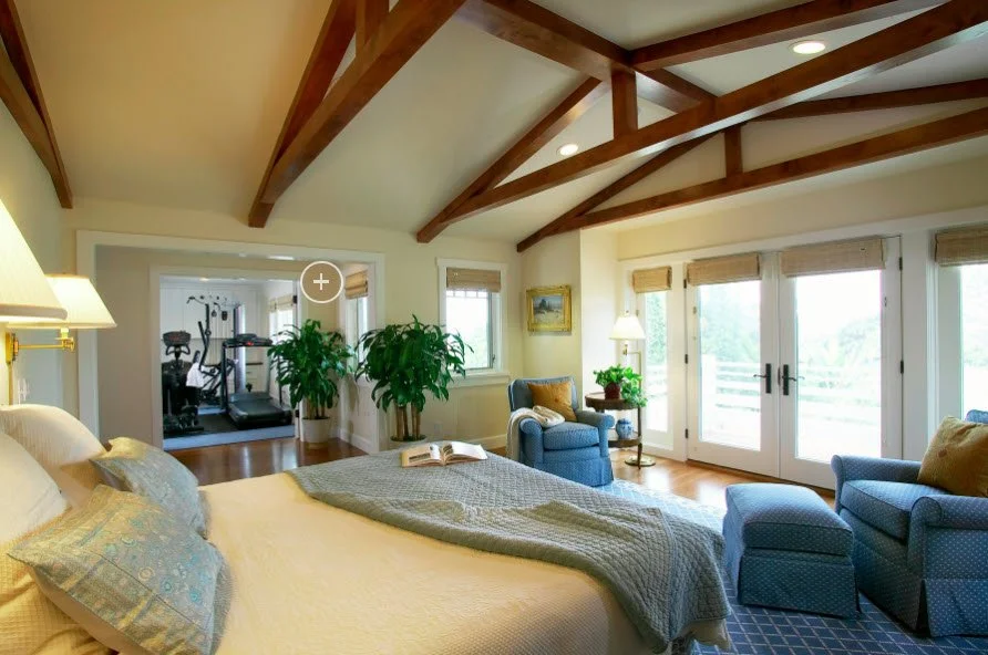 A bright bedroom with a vaulted ceiling and wooden beams, featuring a large bed with pillows and a blanket, a reading chair with an ottoman, a small side table with a lamp, and glass doors leading to a balcony. An exercise area with a treadmill and weights is visible through an open doorway.