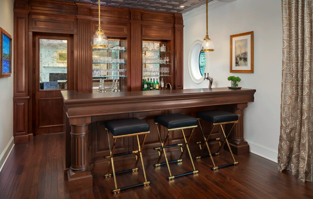 A home bar with a wooden counter and three black and gold bar stools. There are glass shelves with glassware and bottles behind the bar. A small round window and a painting on the white wall are visible. The room has wooden floors, curtains, and hanging pendant lights.
