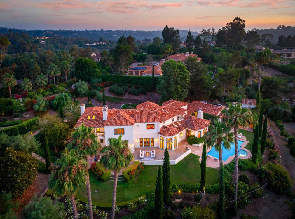 A large, luxurious house with a red tile roof, surrounded by lush greenery and tall palm trees. There is a swimming pool in the backyard, and the house features multiple windows with warm lighting. The landscape includes a variety of trees and bushes, with a scenic view of rolling hills and a colorful sky at sunset.