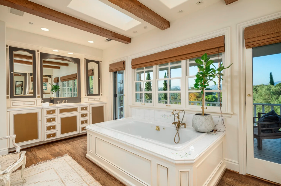 Bright bathroom with a large white bathtub next to multiple windows, a potted plant on the tub, and a view of greenery outside.