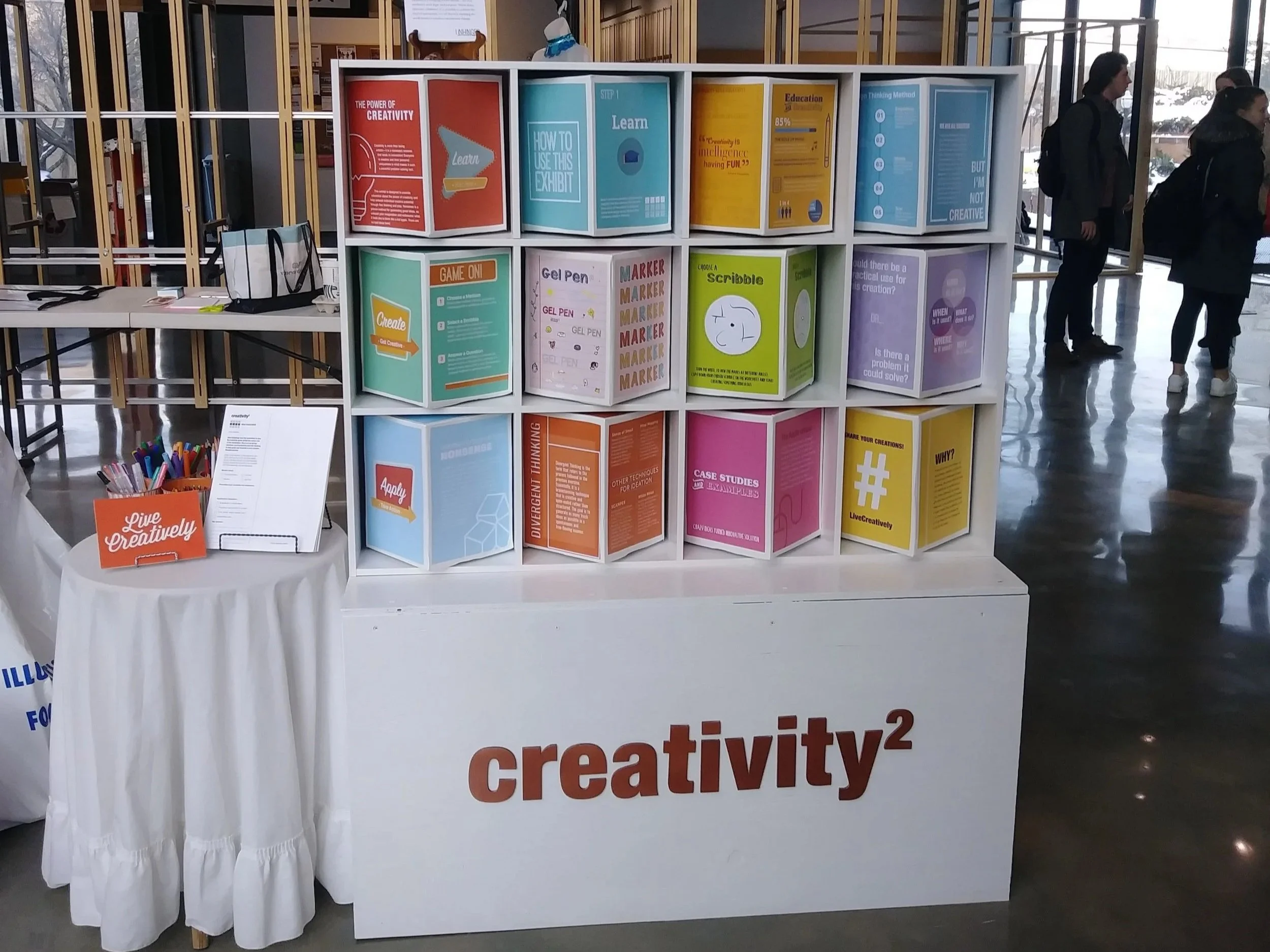 A display booth at an exhibition hall featuring interactive squares and signs about creativity, with several people walking past in the background.