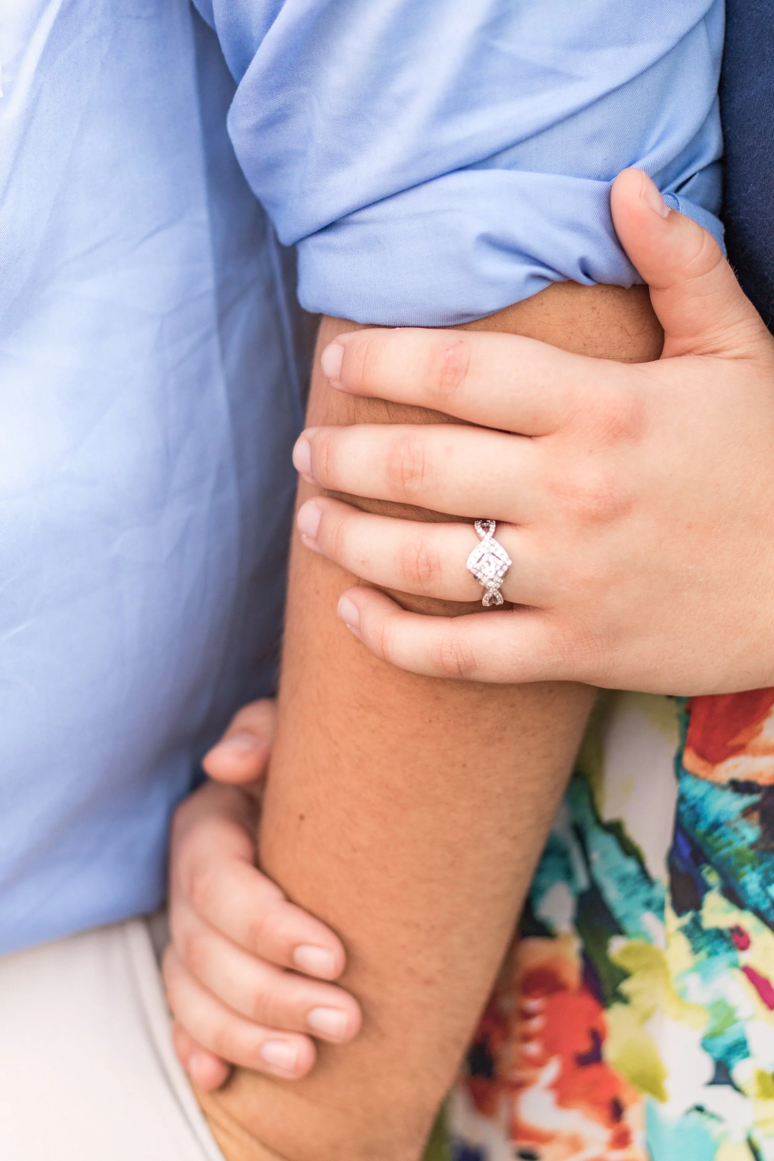Close-up of two people holding each other's arms, one person wearing a blue shirt and the other wearing a colorful floral dress, with a wedding ring visible on the hand of the person in the floral dress.