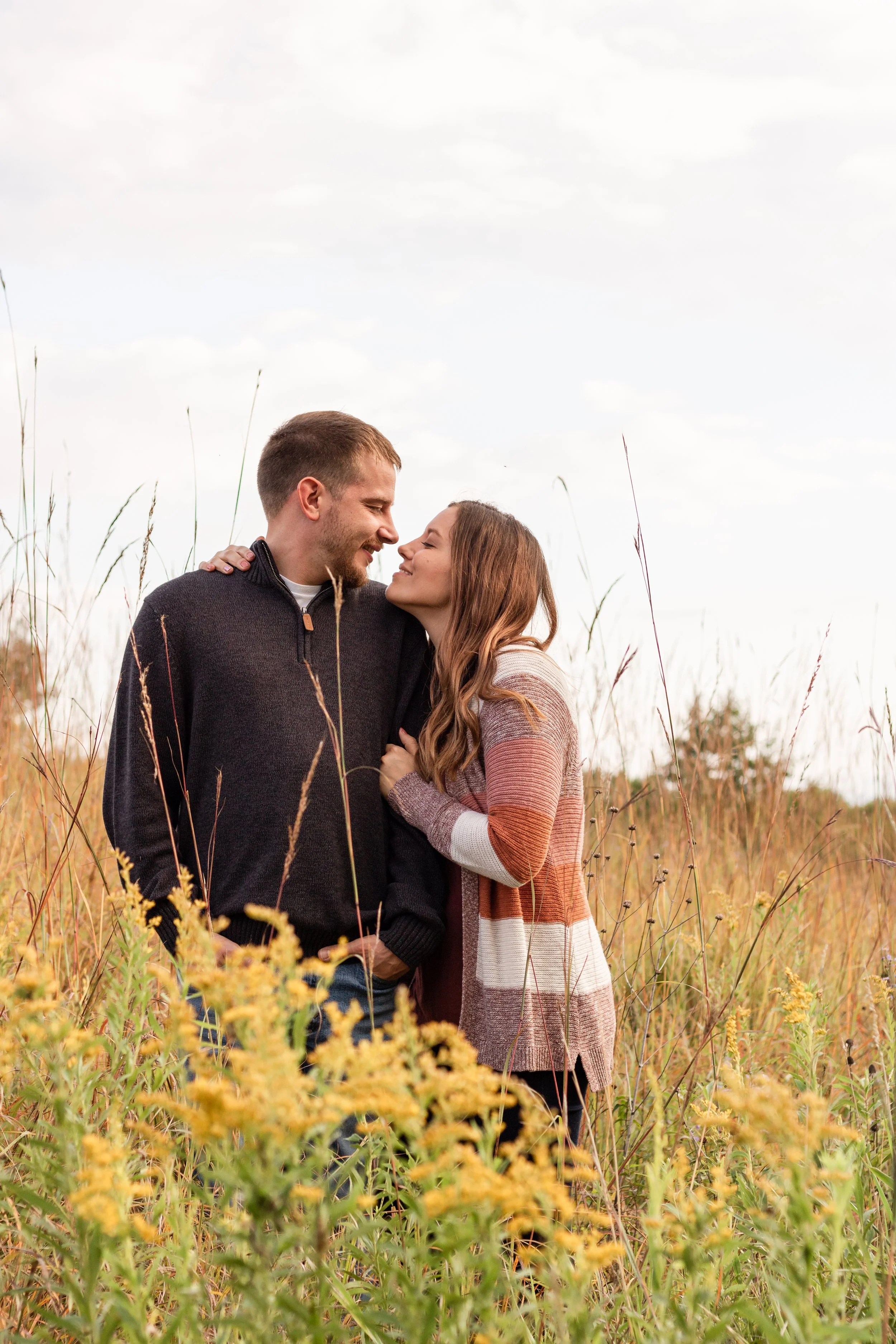 A young couple embracing and smiling in a field with yellow flowers and tall grass, looking into each other's eyes during daytime with clouds in the sky.