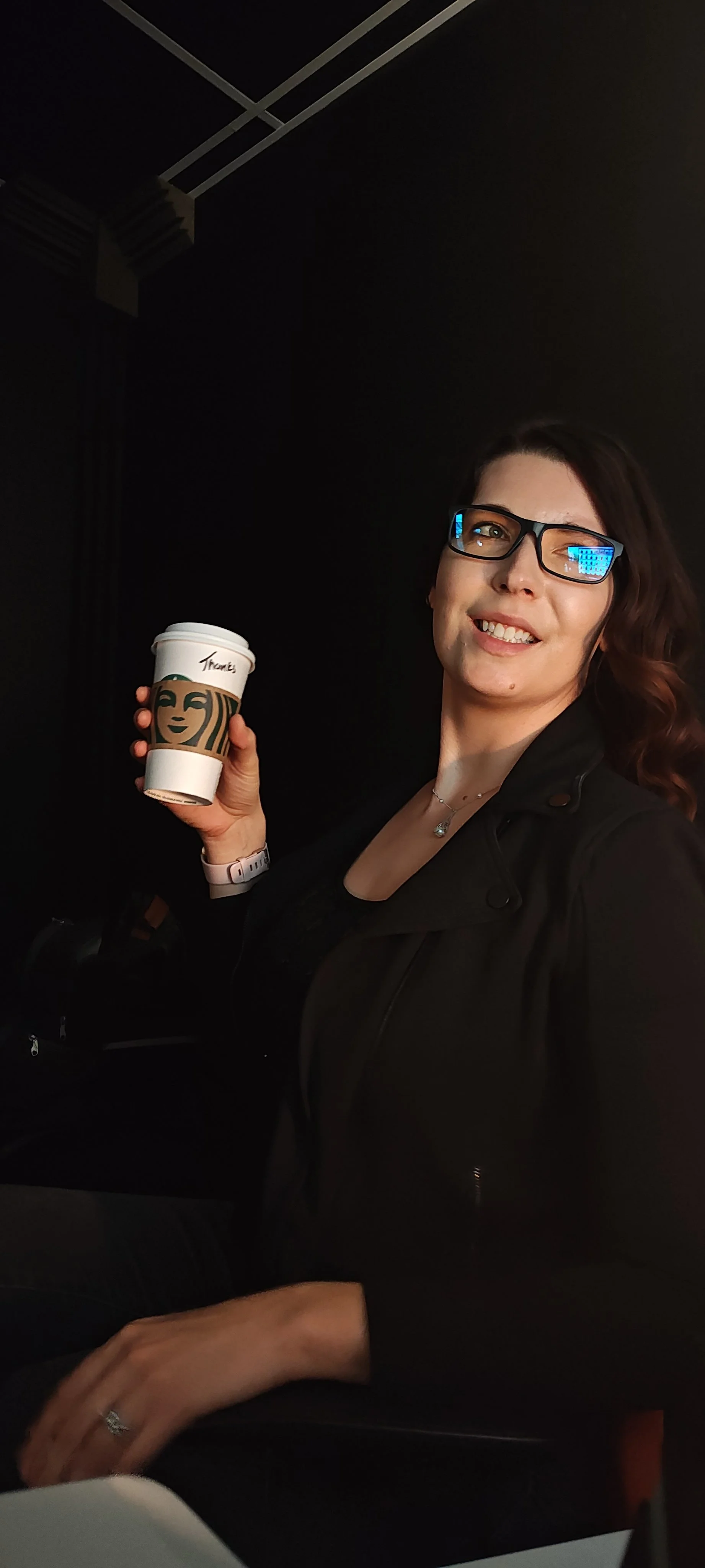 KJ in the dark photo studio, sipping a Starbucks coffee and smiling off-shoulder.