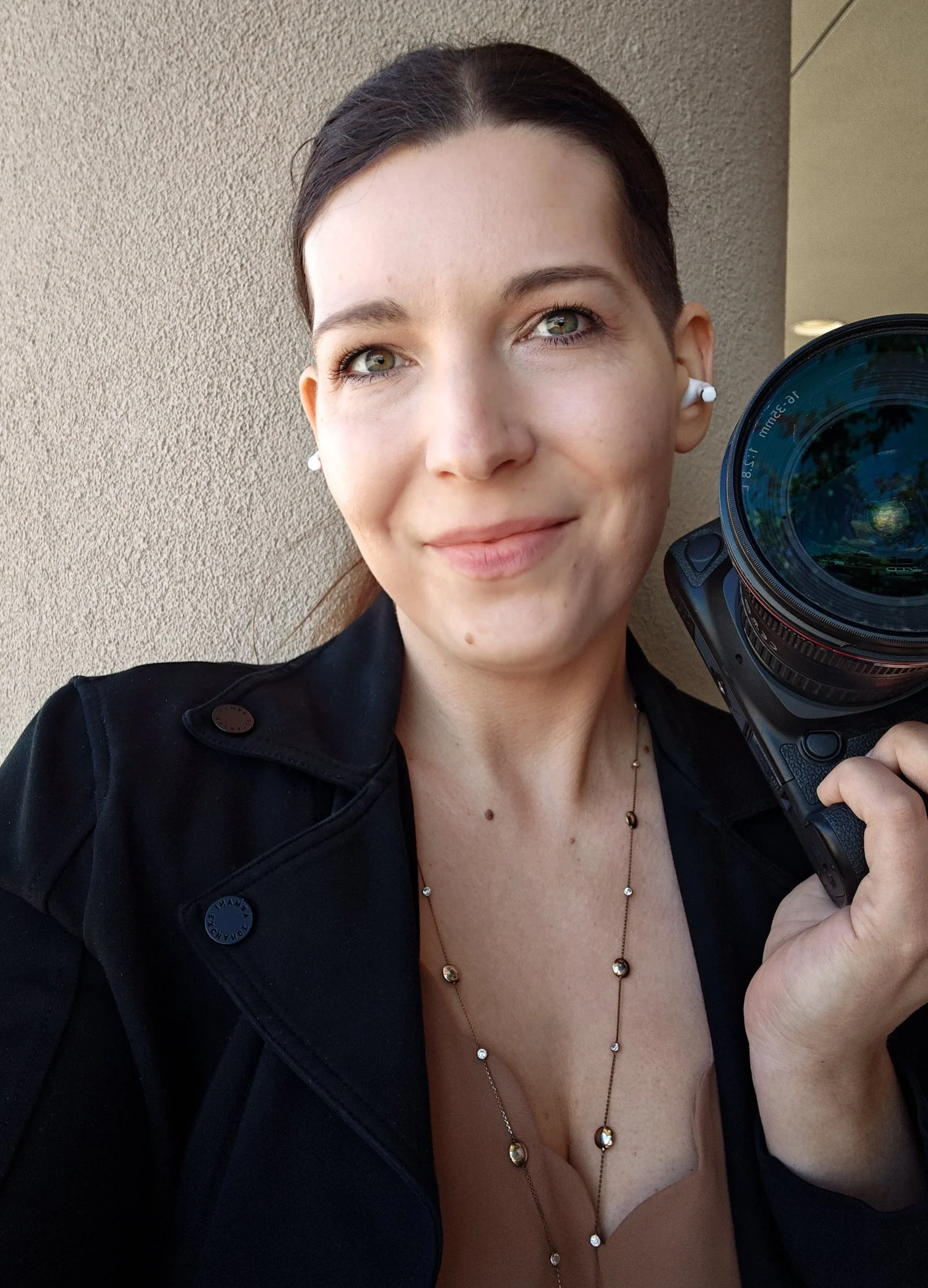 A woman with dark hair, wearing a black jacket, white earbuds, and jewelry, holding a camera, standing against a textured beige wall, smiling slightly.