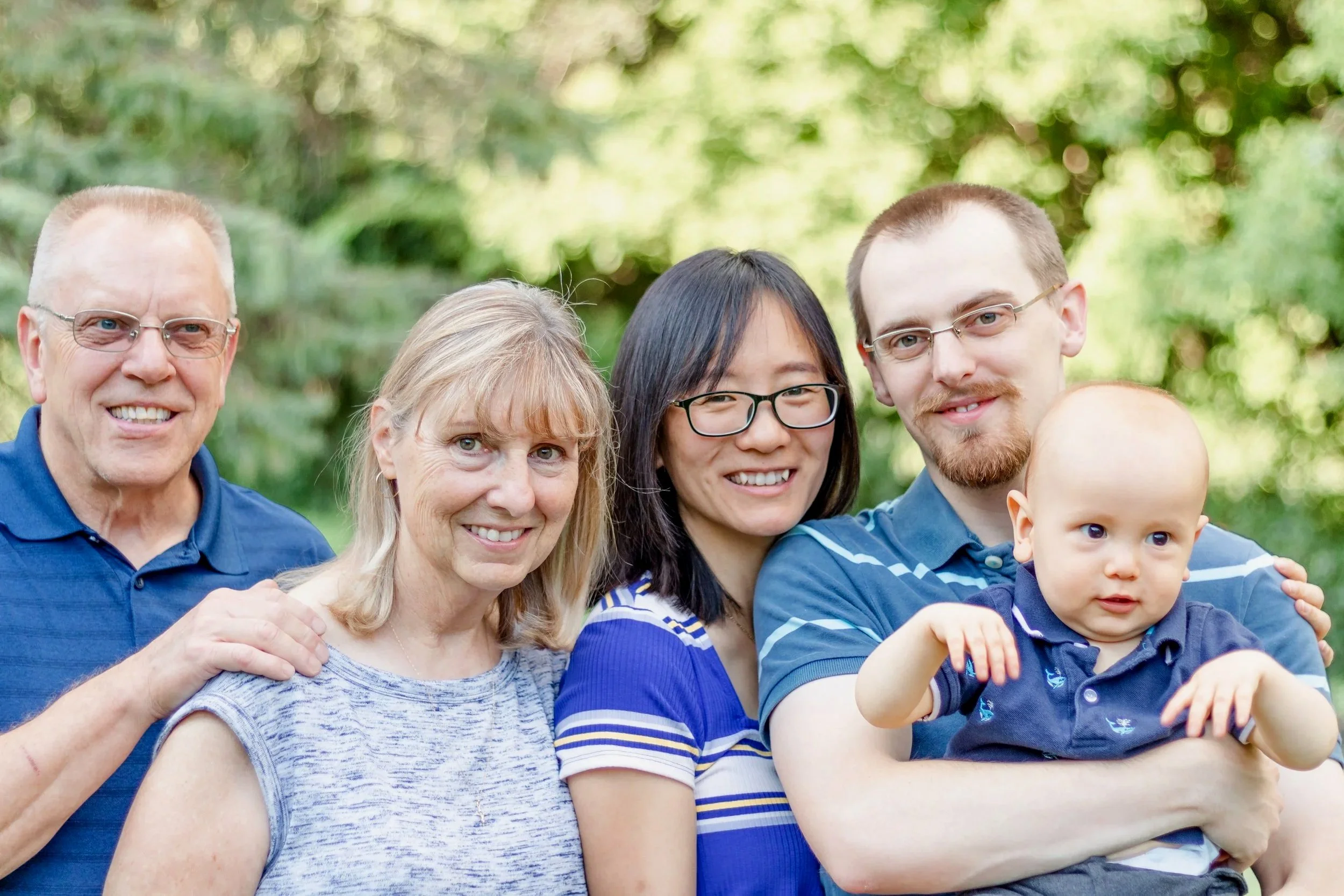 A group of five people, including elderly, middle-aged, and young adults, standing outdoors in front of a blurred green background, smiling and interacting with a baby.