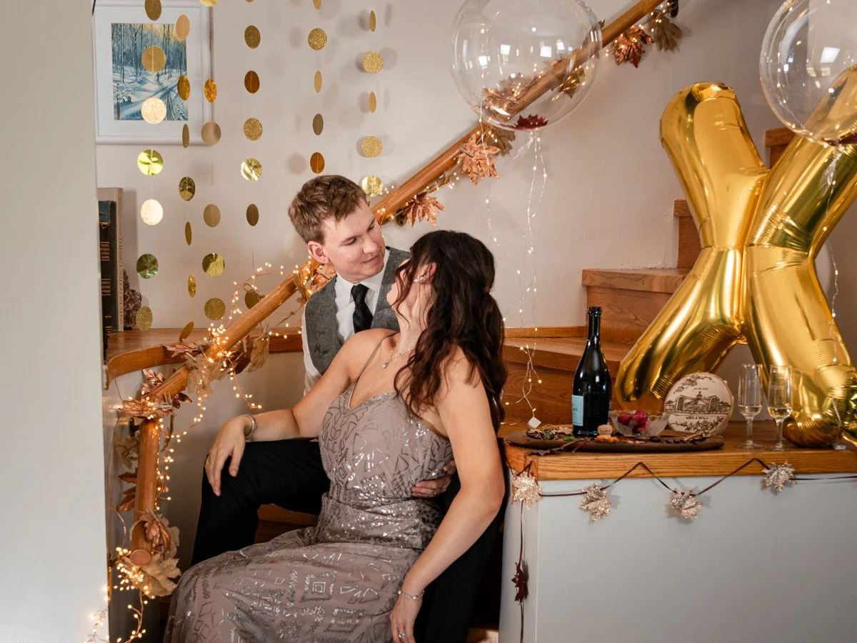 A couple is sitting close together at a decorated indoor party, with gold and clear balloons, fairy lights, and gold leaf garlands. The woman is wearing a sleeveless dress, and the man is dressed in a shirt, tie, and blazer. There are drinks and snacks on the table nearby.