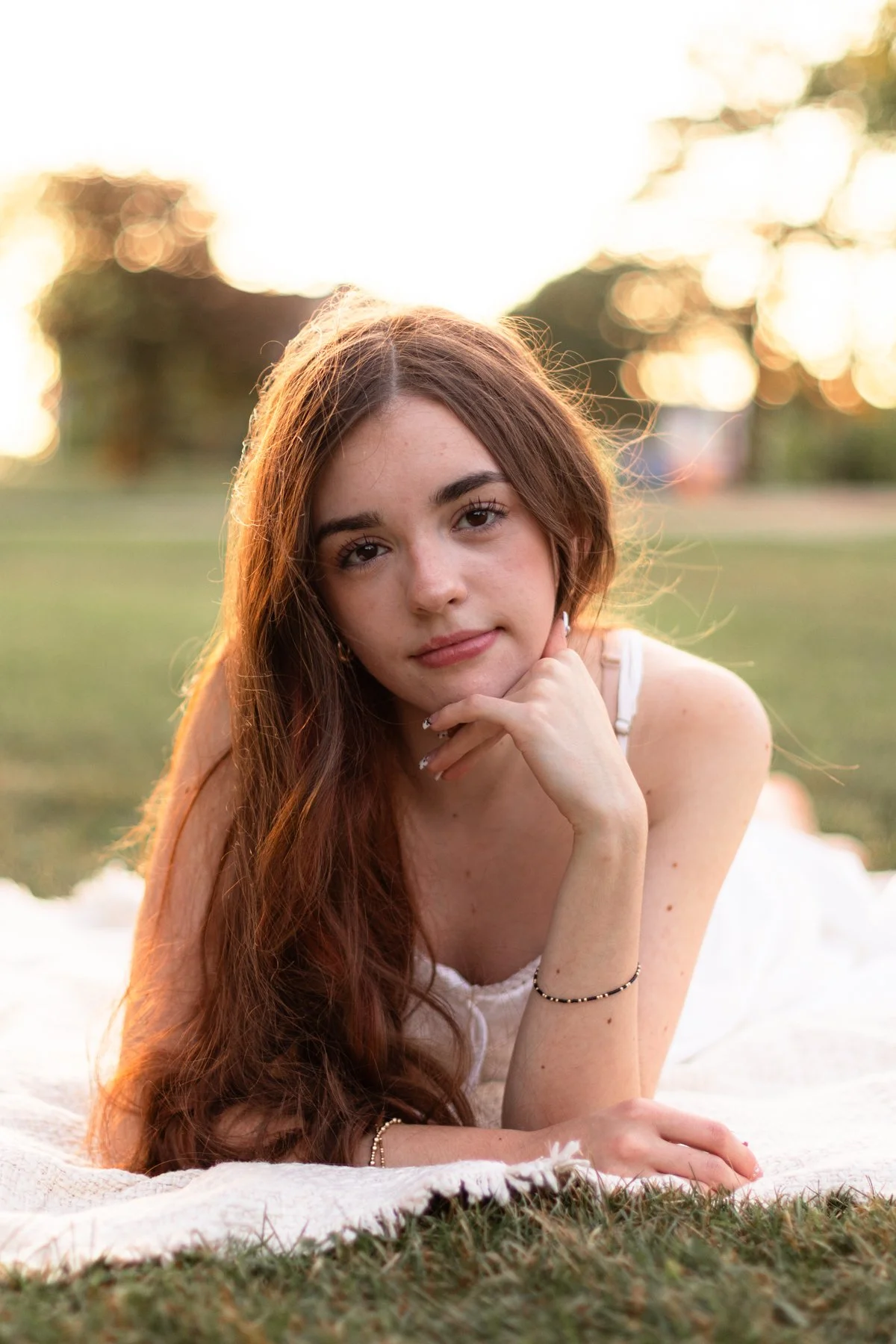 A young woman with long, wavy red hair, lying on a blanket outdoors during sunset, looking at the camera with her chin resting on her hand.