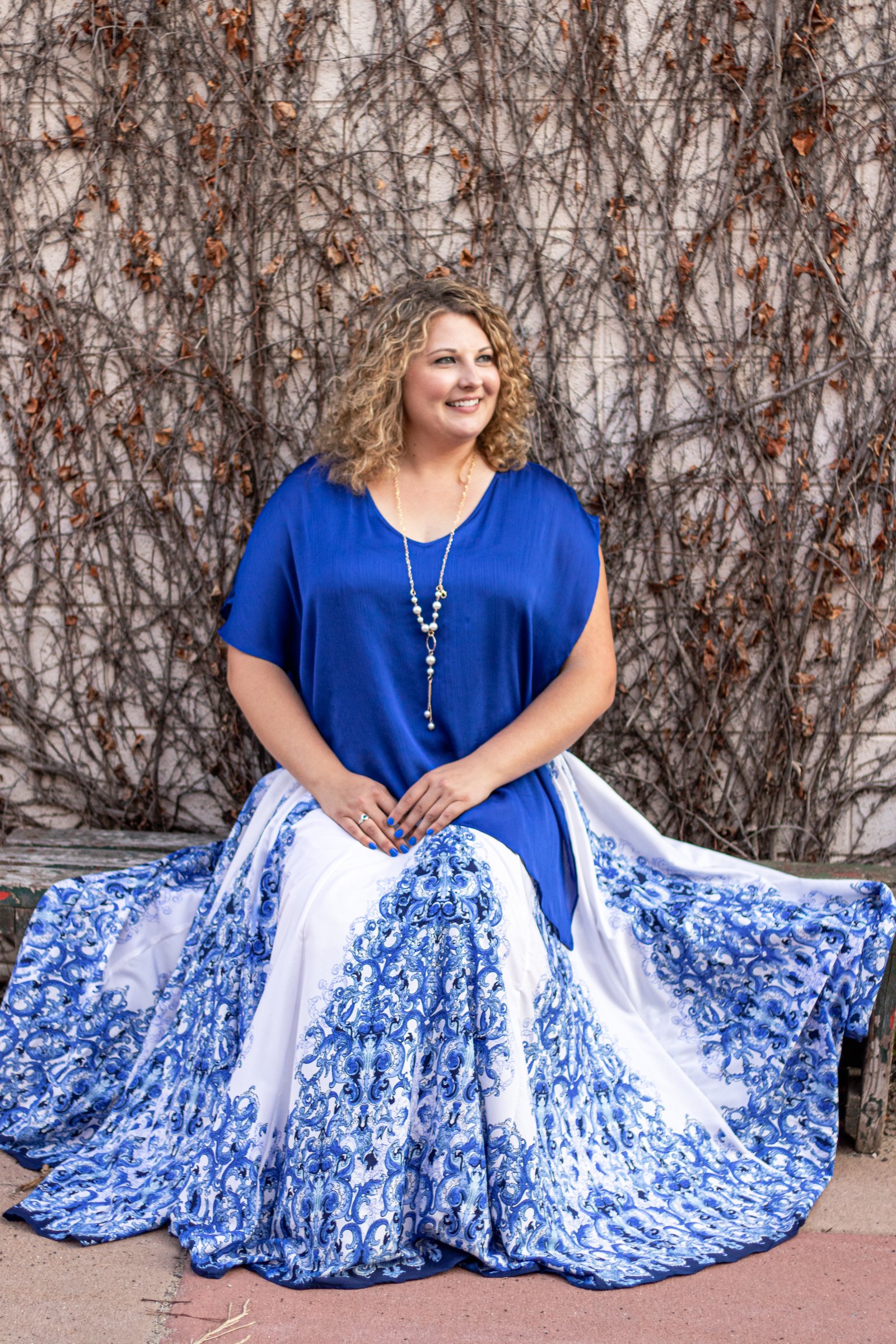 A woman with curly blonde hair sitting in front of a vine-covered wall, wearing a blue top and a flowing white and blue patterned skirt.