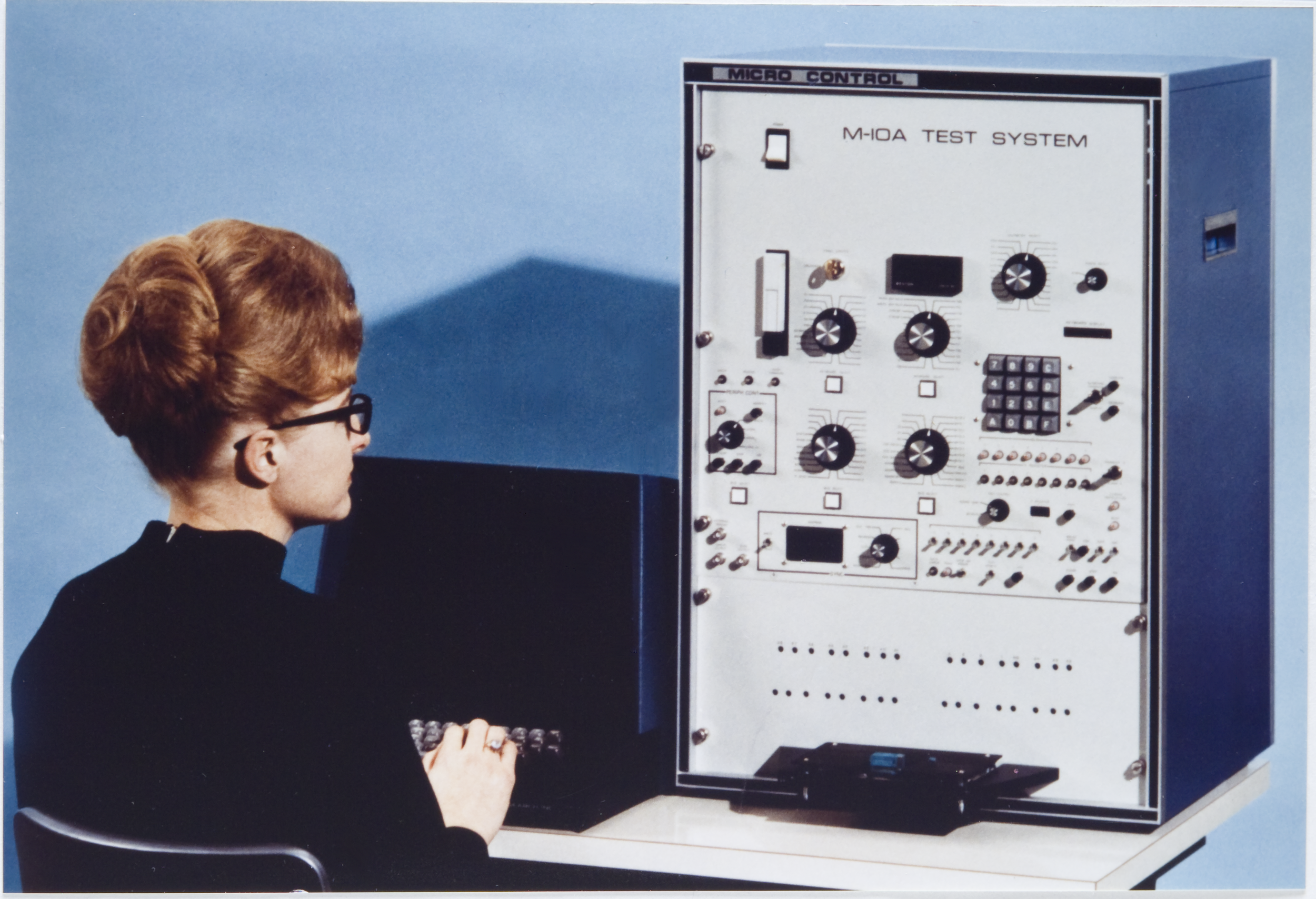 A woman with short red hair, glasses, and wearing a black outfit is operating a vintage machine labeled 'Micro Control' and 'M-IOA Test System' in a room with blue walls.