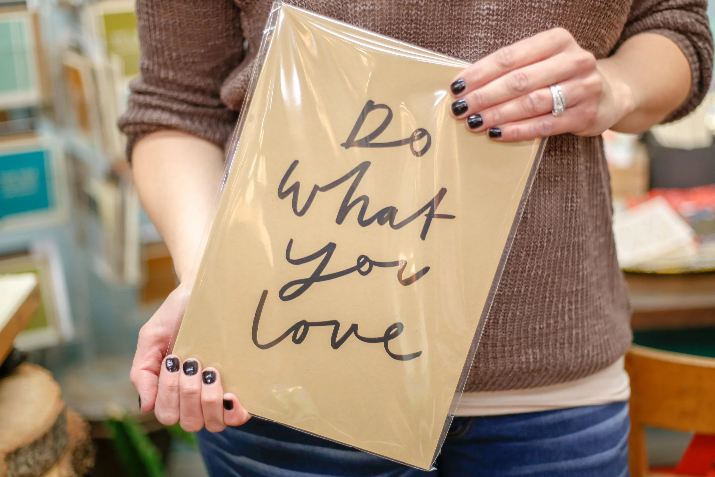 Woman holding sign that says, Do what you love. She has on a brown sweater with black painted nails and blue jeans. The background looks like a retail store even though it is blurry.