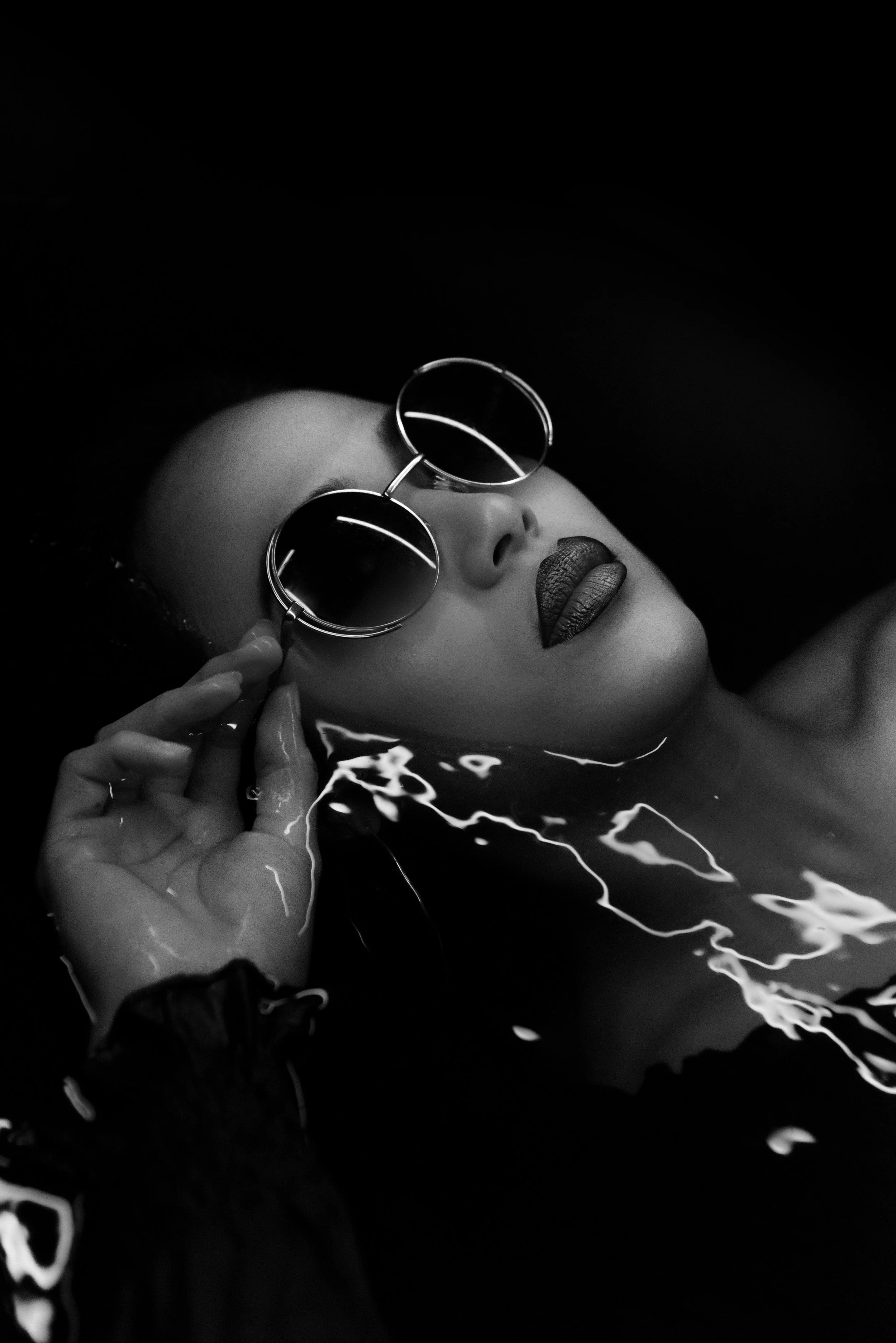 Black and white photo of a woman laying down in the water with her face and part of her hand exposed. She's wearing round sunglasses, lipstick and a black, off the shoulder top.