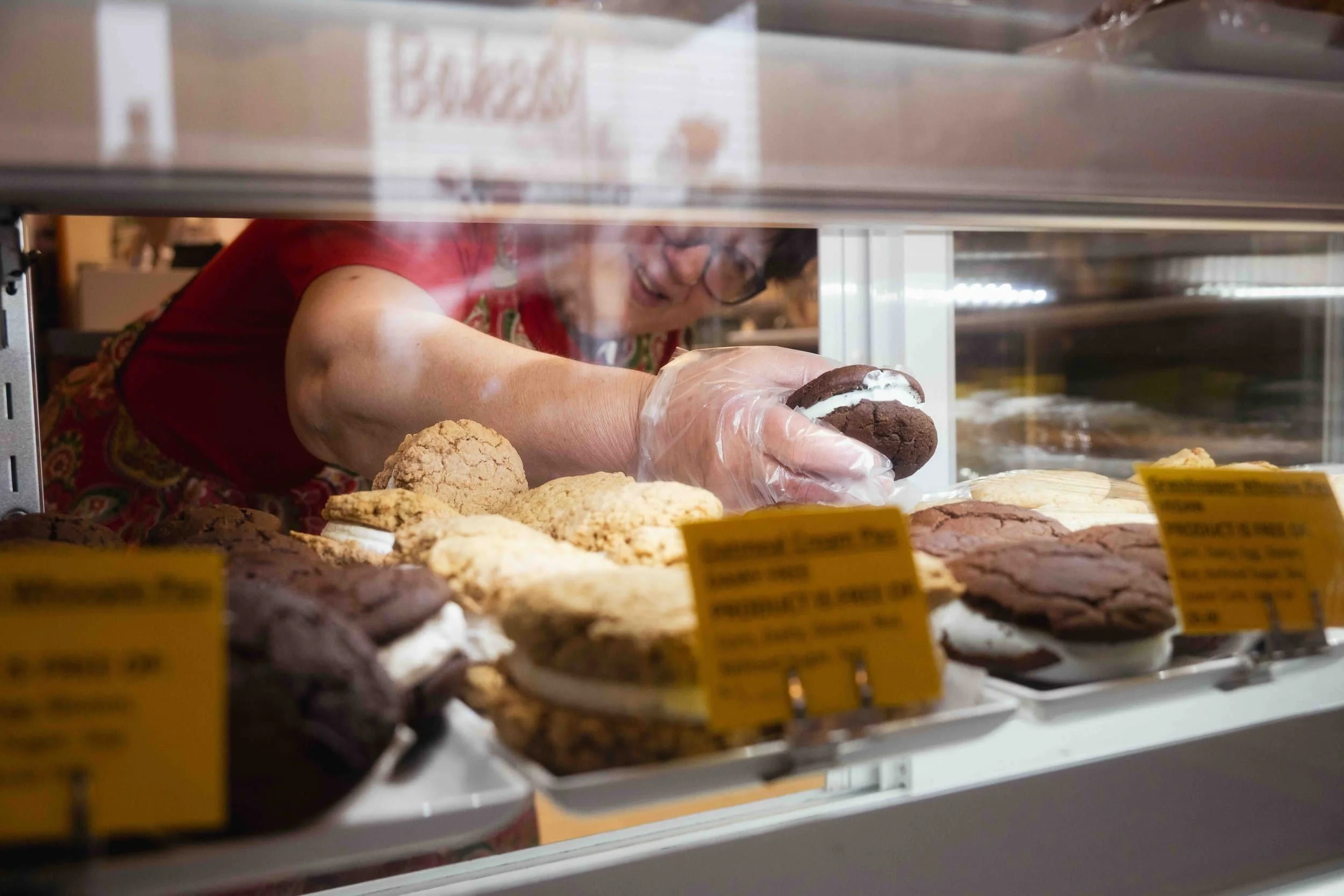A person wearing gloves grabs a whoopie pie from a wholesale bakery display filled with assorted cookie sandwiches.