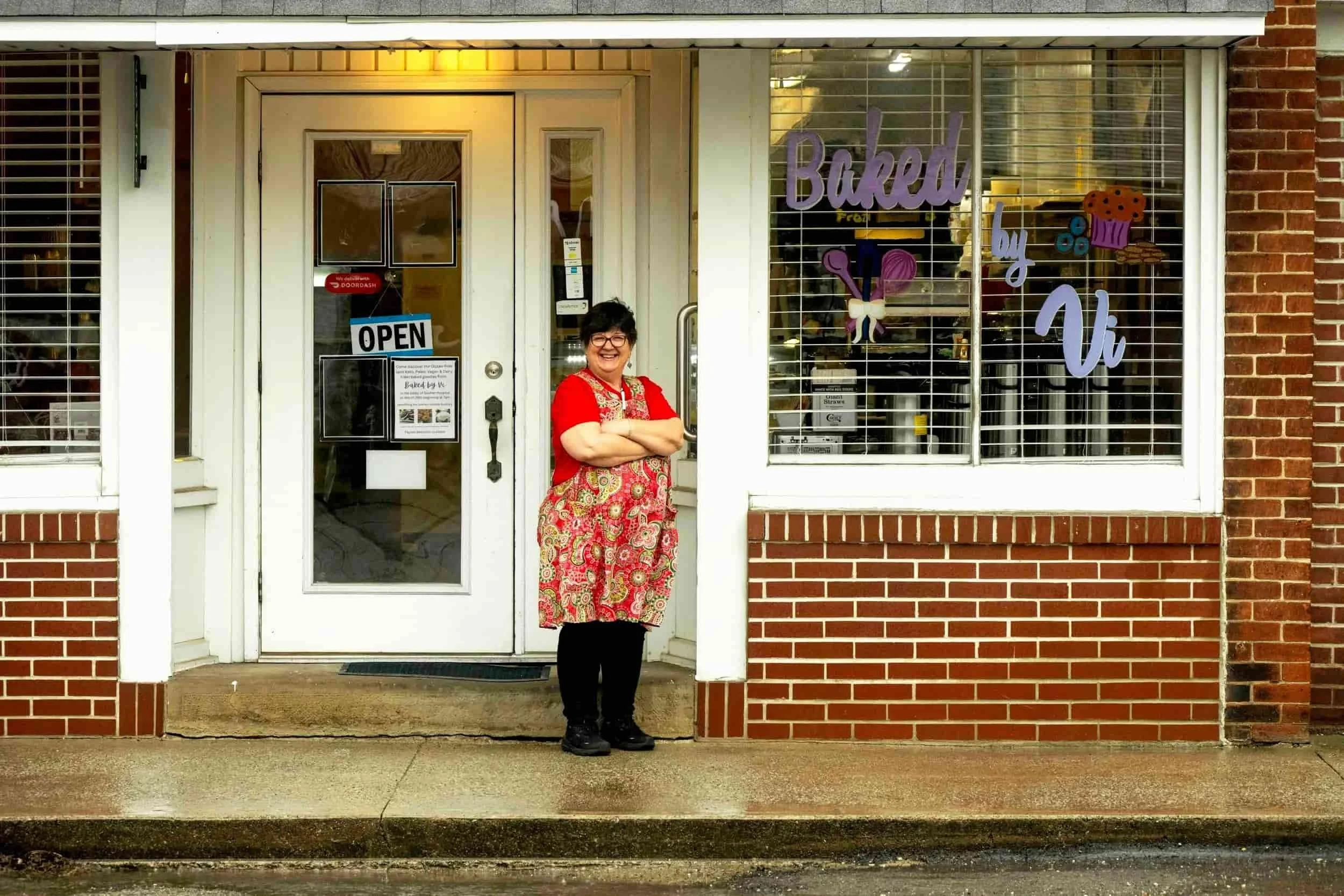 A woman with short dark hair and glasses standing with arms crossed in front of a bakery storefront, wearing a red top and a colorful apron, smiling.