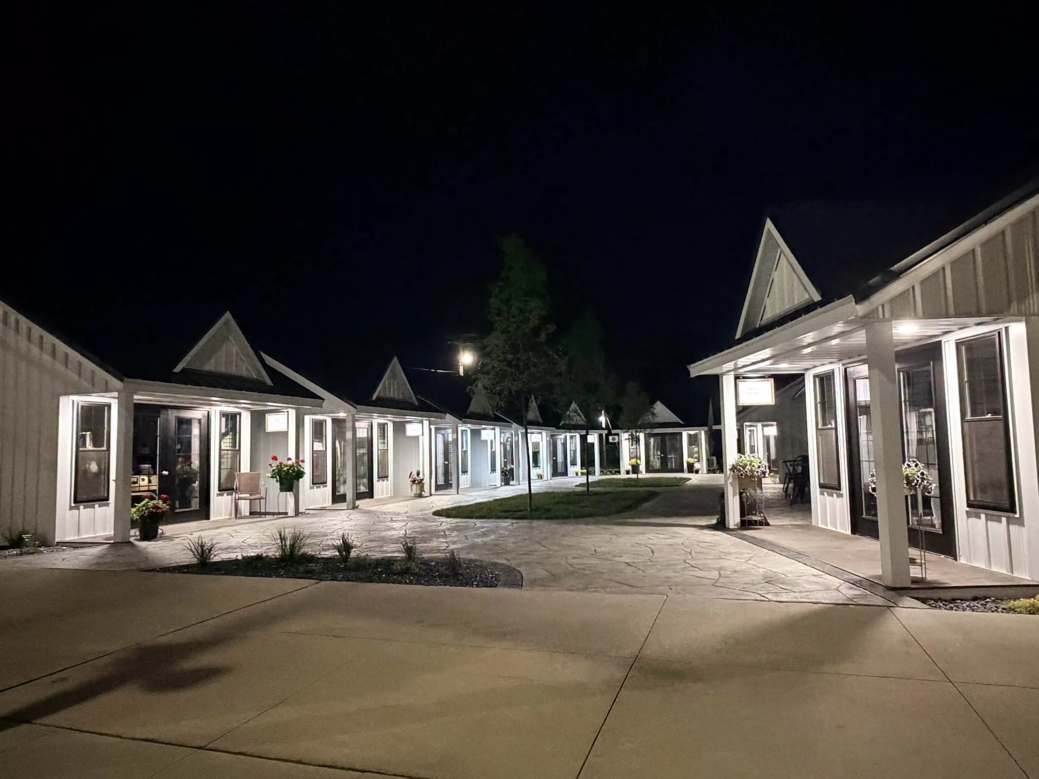 Night view of a row of small, illuminated houses with gabled roofs, surrounded by a paved courtyard, trees, and potted plants.