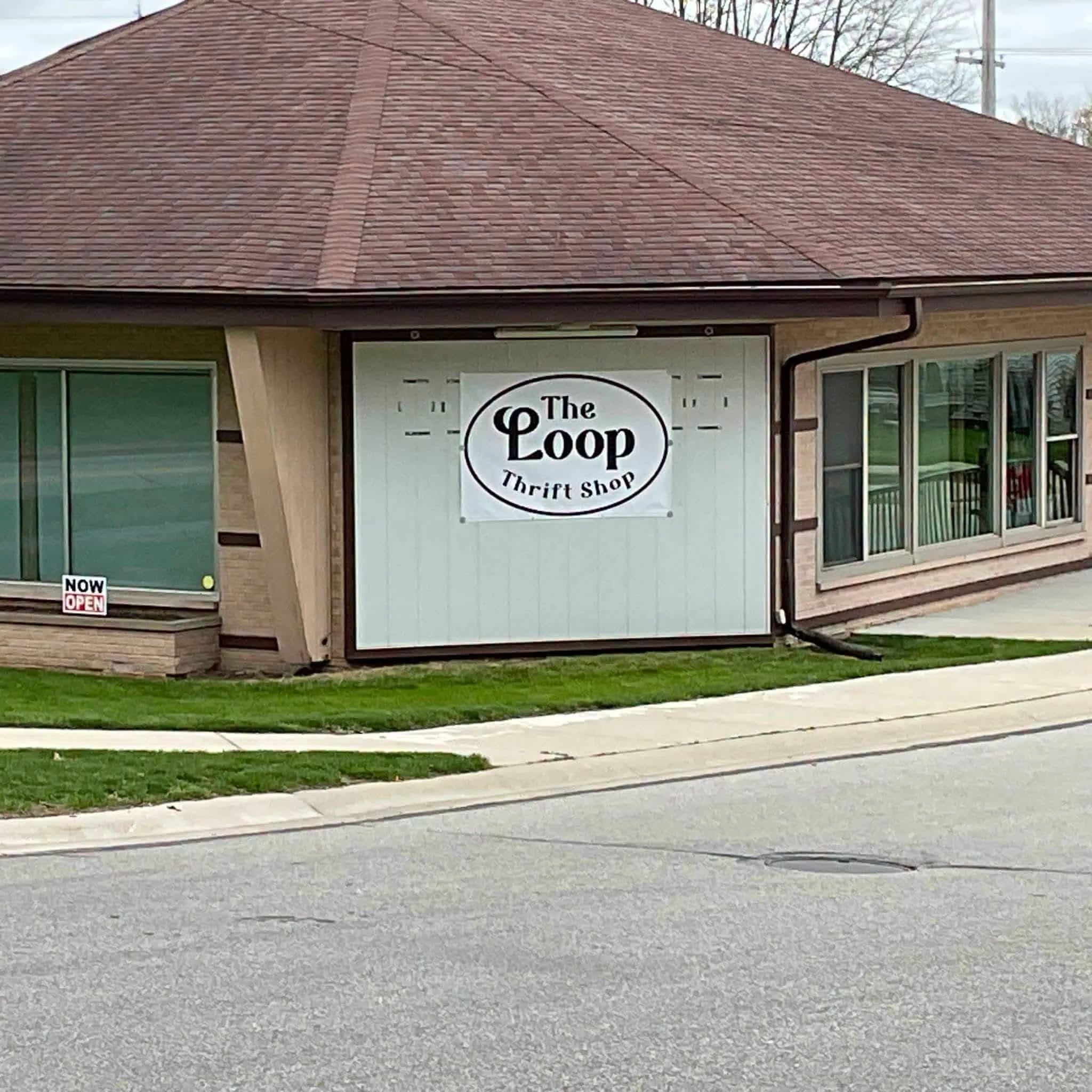 Close-up view of the front of a building with a red shingled roof, beige brick walls, and windows. There is a sign that says "The Loop Thrift Shop" and a small red and white sign that says "Now Open" near the lower-left corner.
