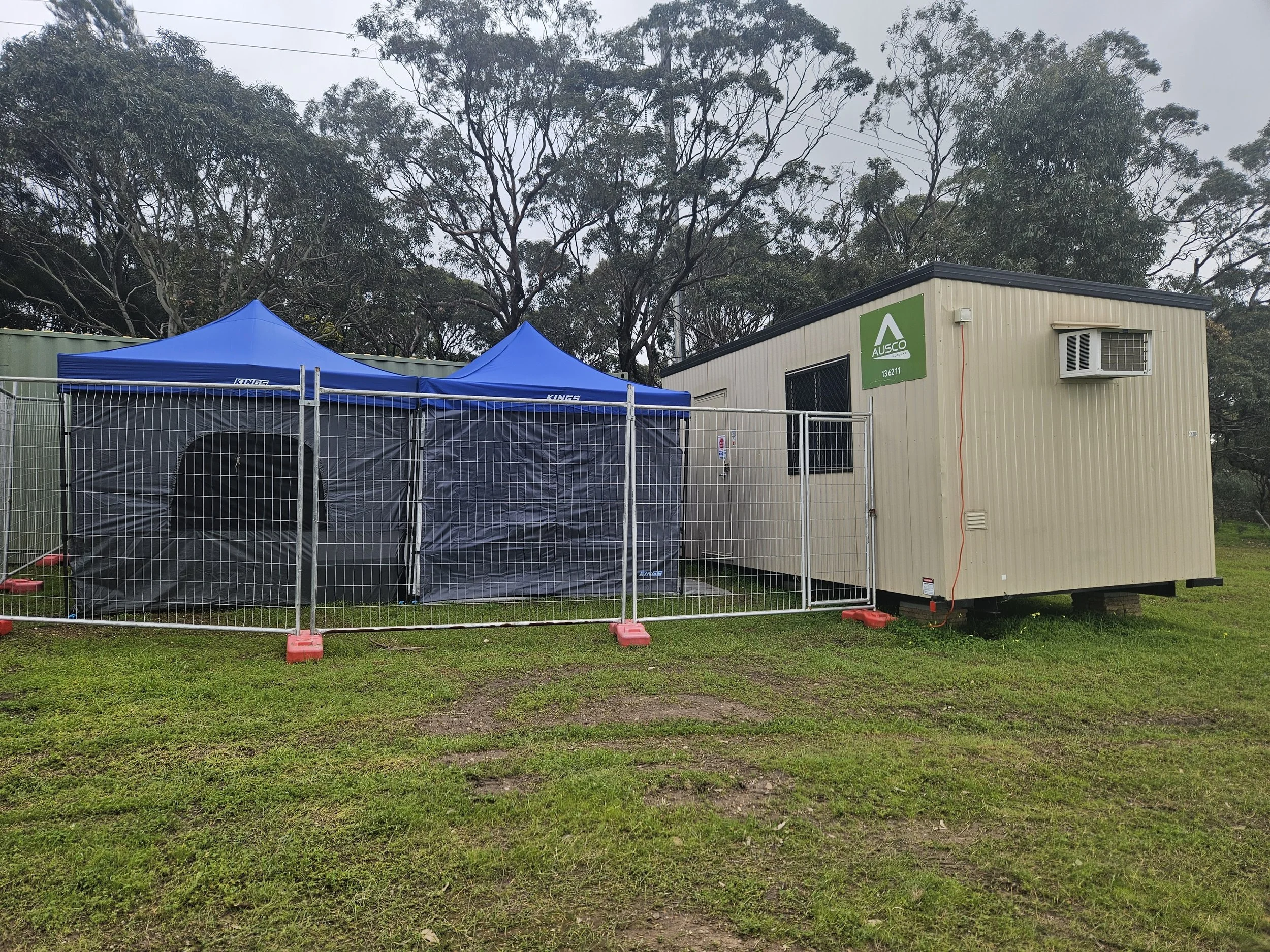 A portable building with an air conditioning unit on the side, along with a green Ausco sign and a metal security door. In front of the building, there are two blue canopies set up, surrounded by a metal fence. The area is outdoors with trees and power lines visible in the background, on a cloudy day.