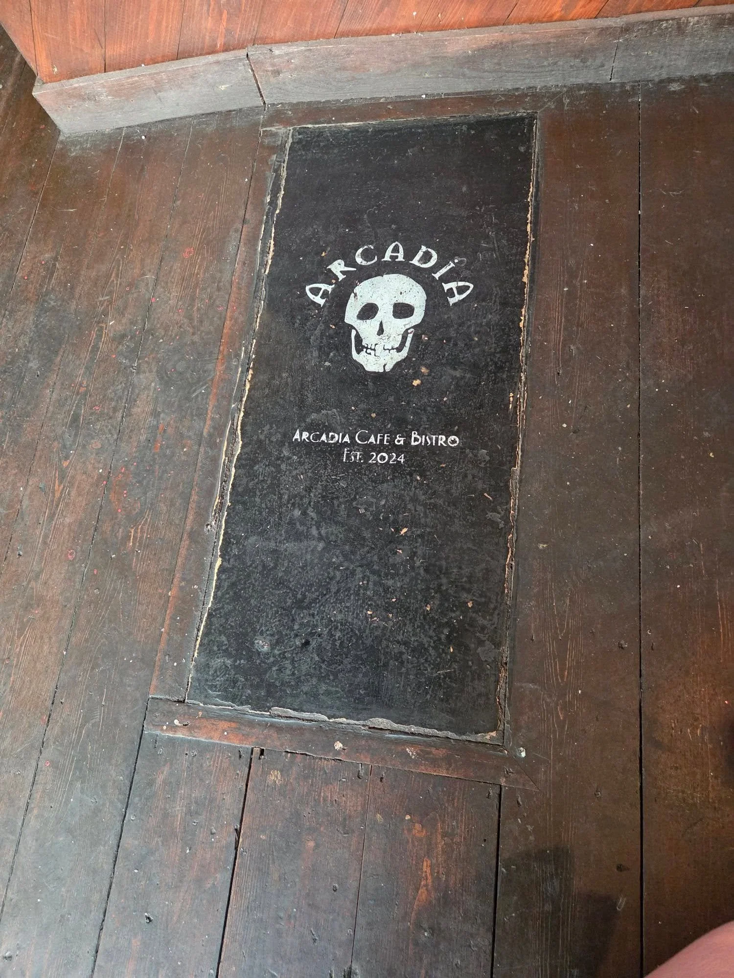 A slab set into wooden flooring, meant to look like a grave, with the name of the establishment Arcadia Cafe and Bistro and the date 2024 inscribed into it