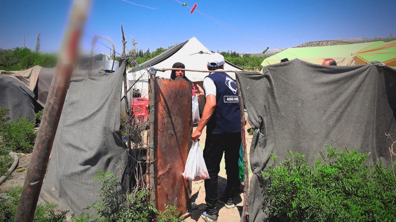 Une personne avec un gilet d'association humanitaire portant des sacs en plastique entre dans un camp de réfugiés avec des tentes et des abris de fortune entourés de végétation, sous un ciel bleu.