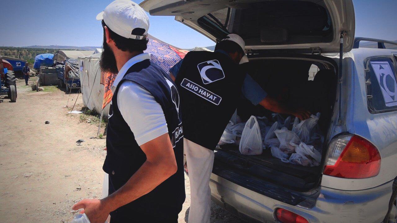 Des bénévoles distribuent des sacs de nourriture à partir du coffre d'une voiture dans un camp. Une tente et un tracteur sont visibles en arrière-plan.