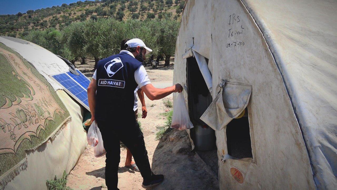 Un homme en uniforme distribue des vivres à l'entrée d'une tente dans un camp, avec des panneaux solaires et des arbres à l'arrière-plan.