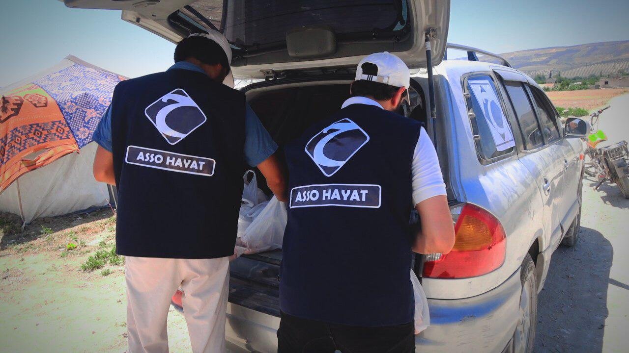 Deux personnes portant des gilets identifiables, avec le logo "Asso Hayat", chargent des articles dans le coffre d'une voiture grise garée près d'un campement de tentes.