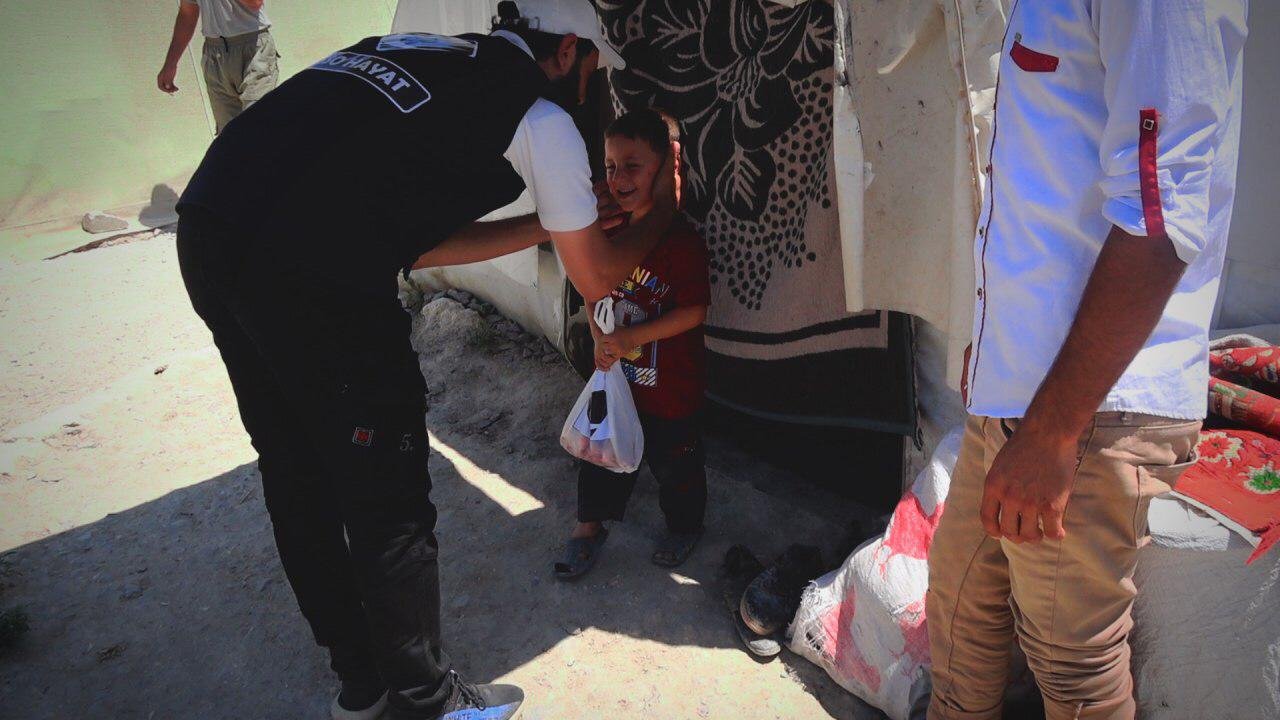 Un homme réconforte un enfant tenant un sac en plastique devant une tente, avec d'autres personnes autour.