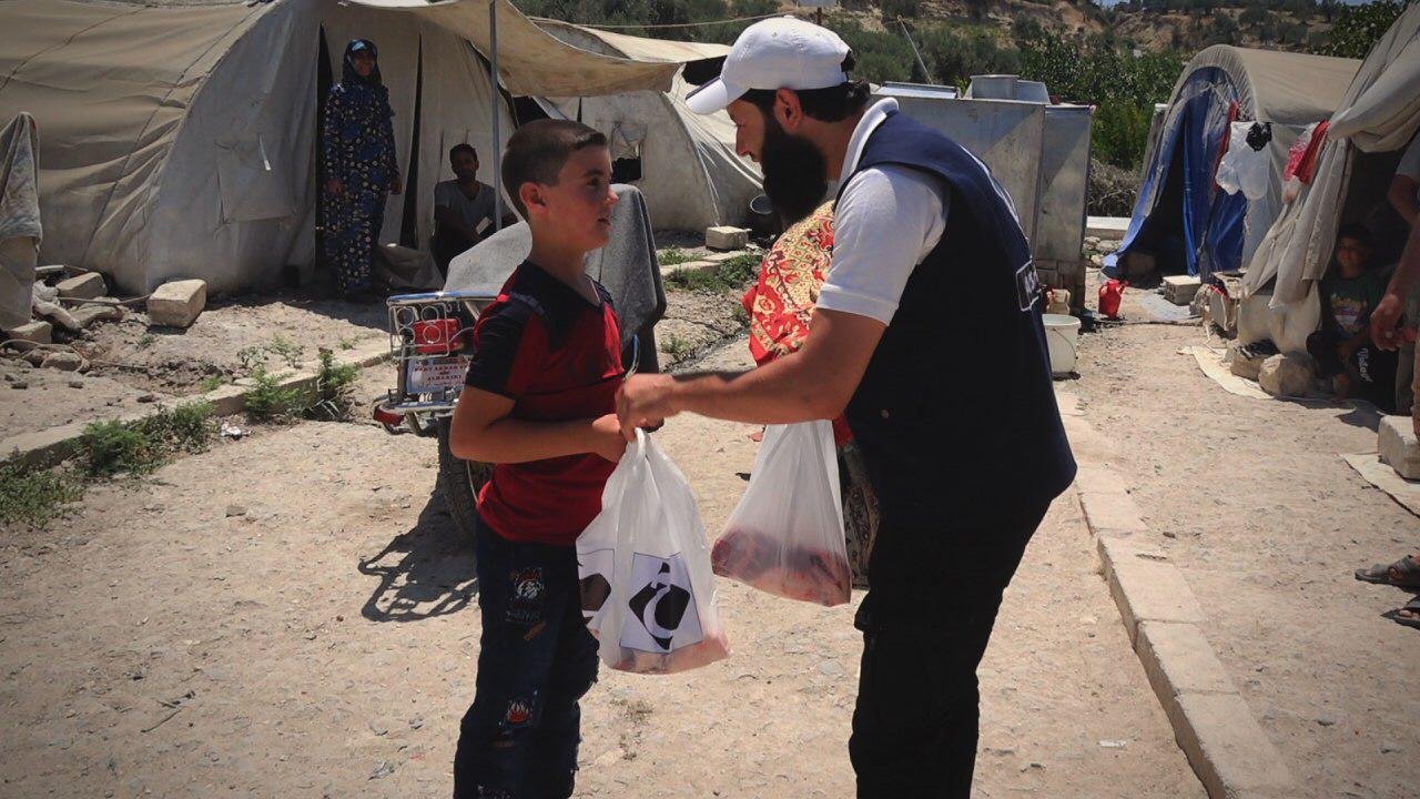 Un homme remettant des sacs à un garçon, près de tentes de réfugiés, avec d'autres personnes à l'arrière-plan.
