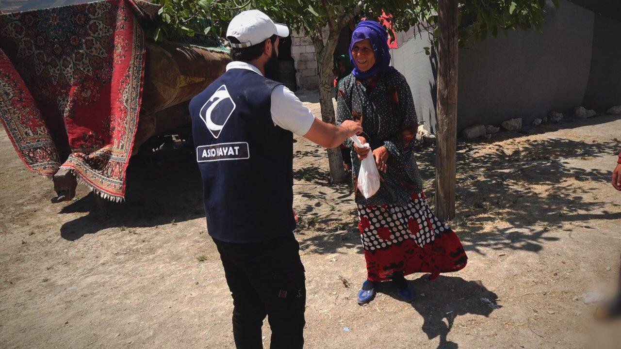 Un homme en gilet avec logo "ASIO HAYAT" donne un sac à une femme portant un foulard violet et une jupe colorée. Ils sont à l'extérieur près d'un arbre et d'une charrette couverte de tapis.