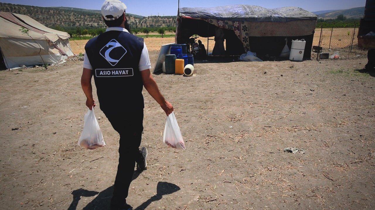 Un homme en uniforme portant deux sacs plastiques marche vers une tente dans un campement en plein air avec des collines en arrière-plan.