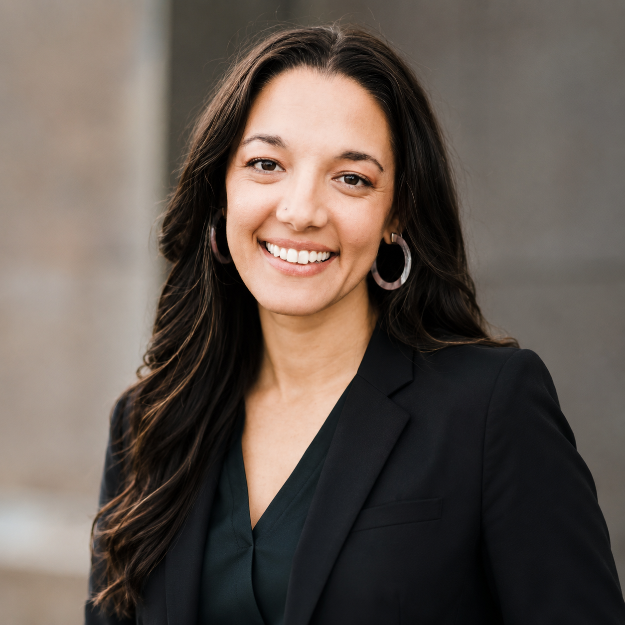 Woman with brown wavy hair, brown eyes with large pink hoop earrings smiling with black blouse and blazer on.