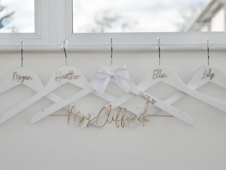 Five white wooden wedding hangers hanging in a row, featuring custom handmade gold wire names including "Mrs Clifford" with a white bow, "Megan", "Heather", "Ellie", and "Lily" for a bridal party.