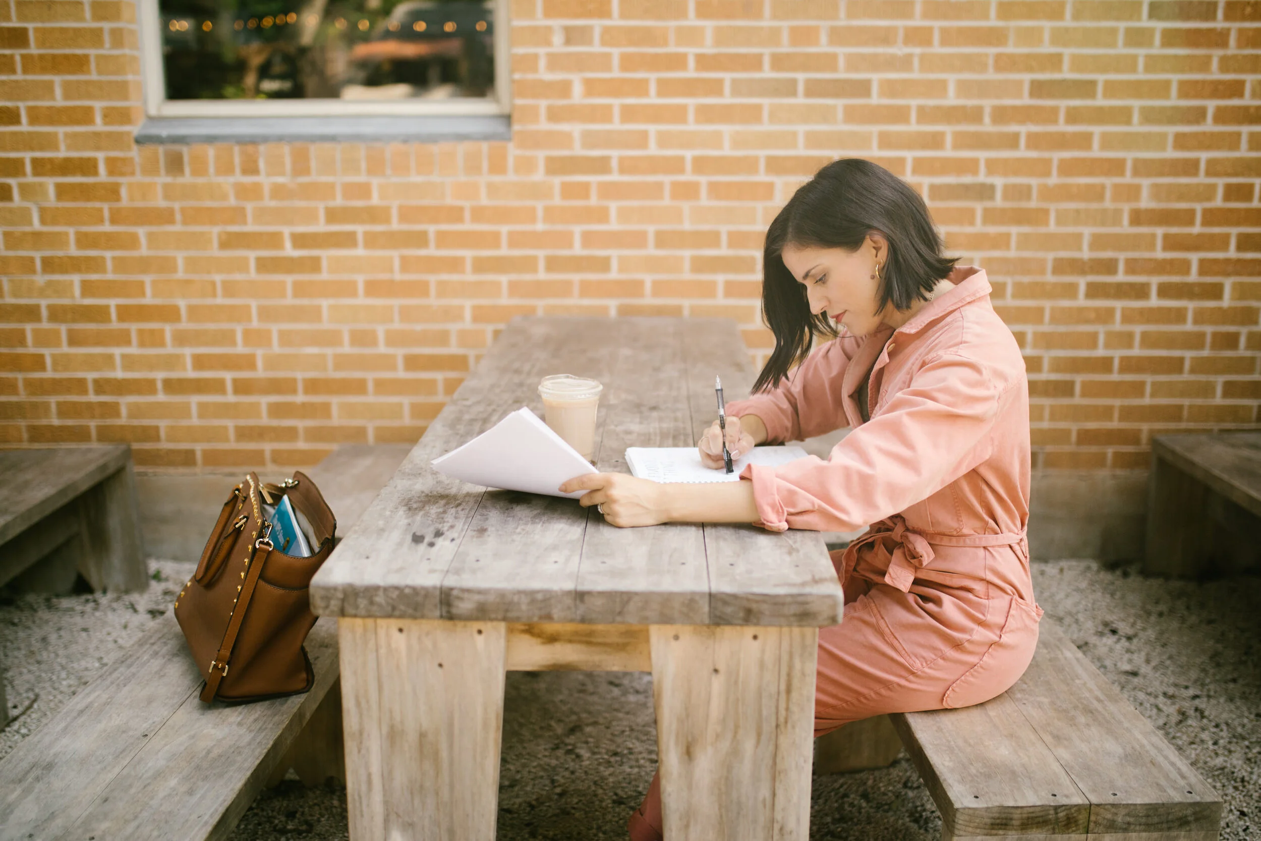 A woman sitting at a wooden outdoor table with a brick wall behind her, writing in a notebook with a pen. There is a coffee drink next to her and a brown handbag on the bench.