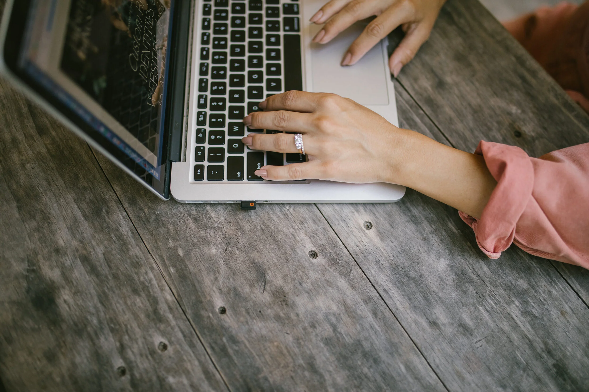 A person typing on a silver laptop with a black keyboard, wearing a wedding ring, seated at a rustic wooden table.
