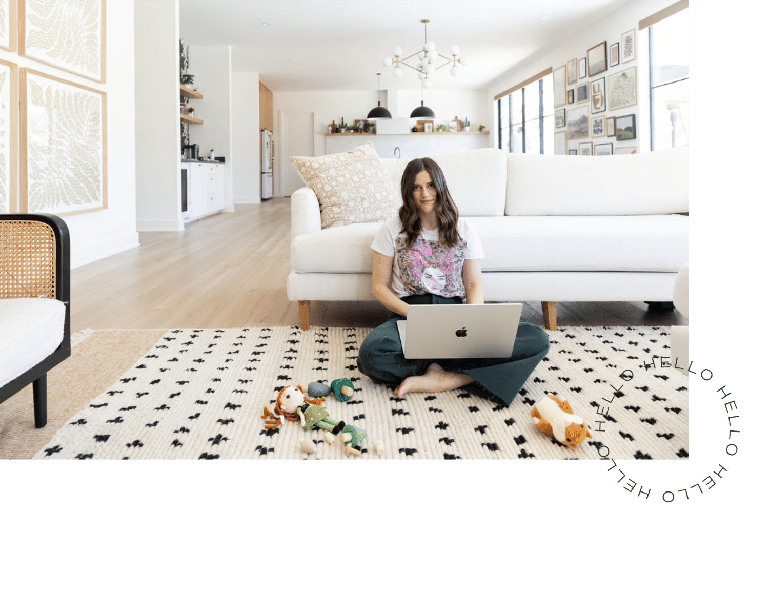 A young woman sitting on a black and white patterned rug with a silver Apple laptop on her lap in a bright, modern living room. She is smiling, with toys scattered on the rug around her, and a white couch and gallery wall of framed art in the background.