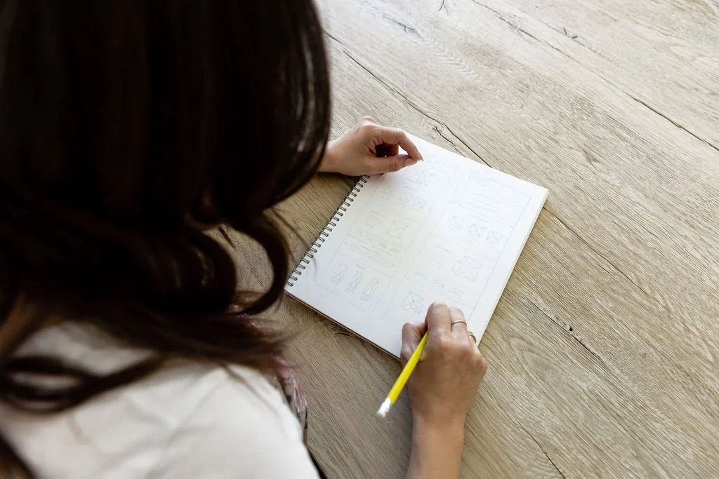 A woman with dark hair writing in a spiral notebook with a yellow pencil on a wooden table.
