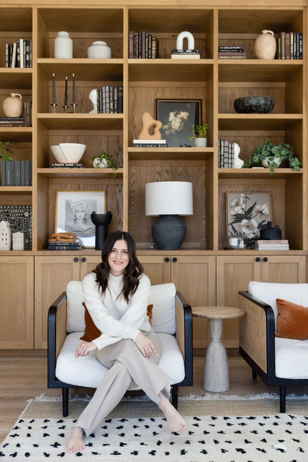 A woman sitting on a white cushioned chair in a living room with a large wooden bookshelf behind her, decorated with books, art, and vases. The room has a modern, minimalist style with neutral tones.