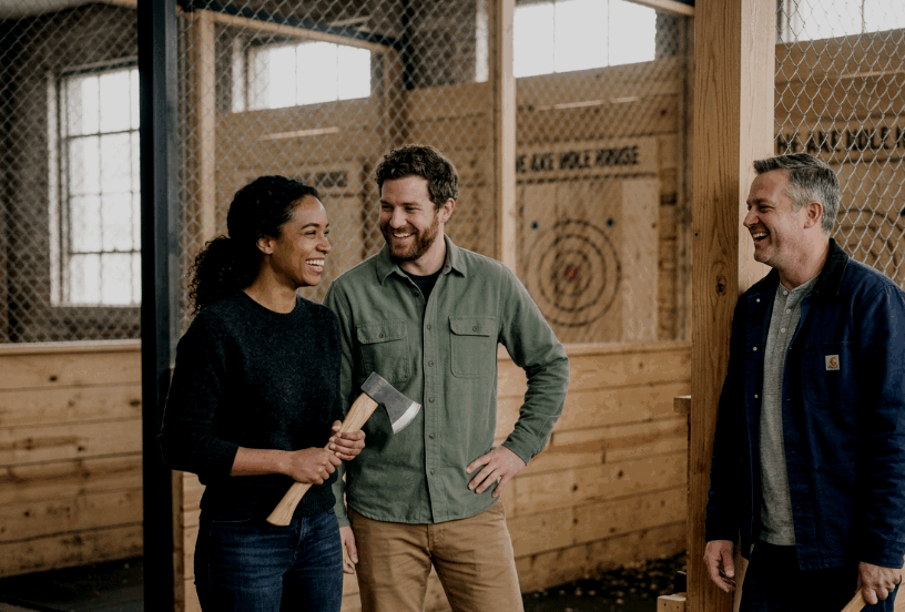 Three people, two men and a woman, smiling and talking inside a wooden axe-throwing venue. The woman holds an axe.