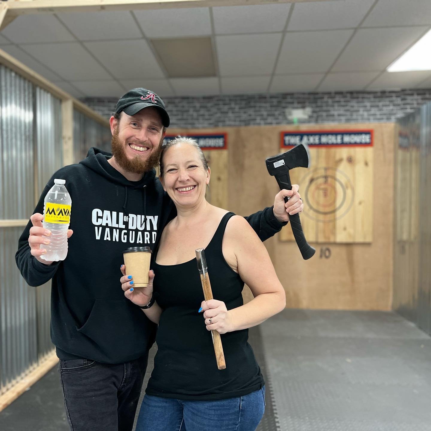 A man and woman smiling at an axe-throwing venue, with the man holding a water bottle and a woman holding a drink, both standing in front of a target.